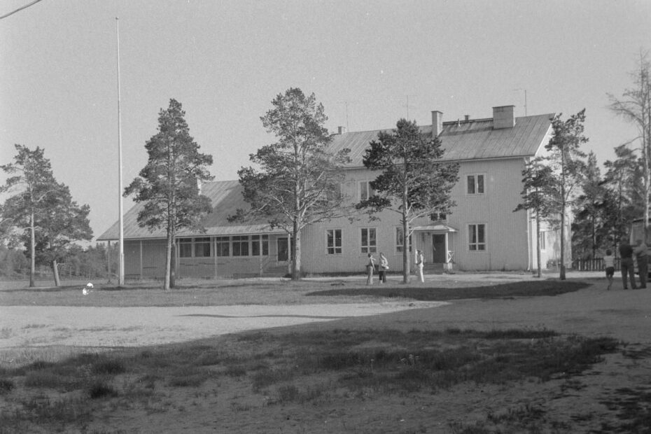 Mustavalkoisessa kuvassa kaksikerroksinen vaalea rakennus, jonka edessä kolme mäntyä. Yhden männyn luona kolme ihmistä seisoo keskustelemassa.