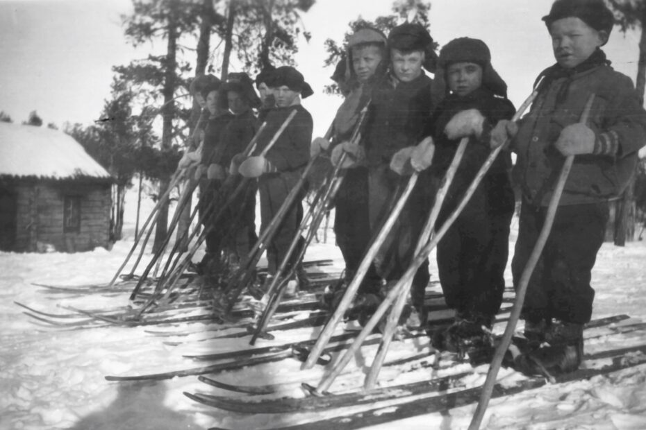 Mustavalkoisessa kuvassa ryhmä poikia rinnakkain puusukset jalassa ja puiset sauvat kädessä. Taustalla yksikerroksinen hirsirakennus ja mäntyjä.